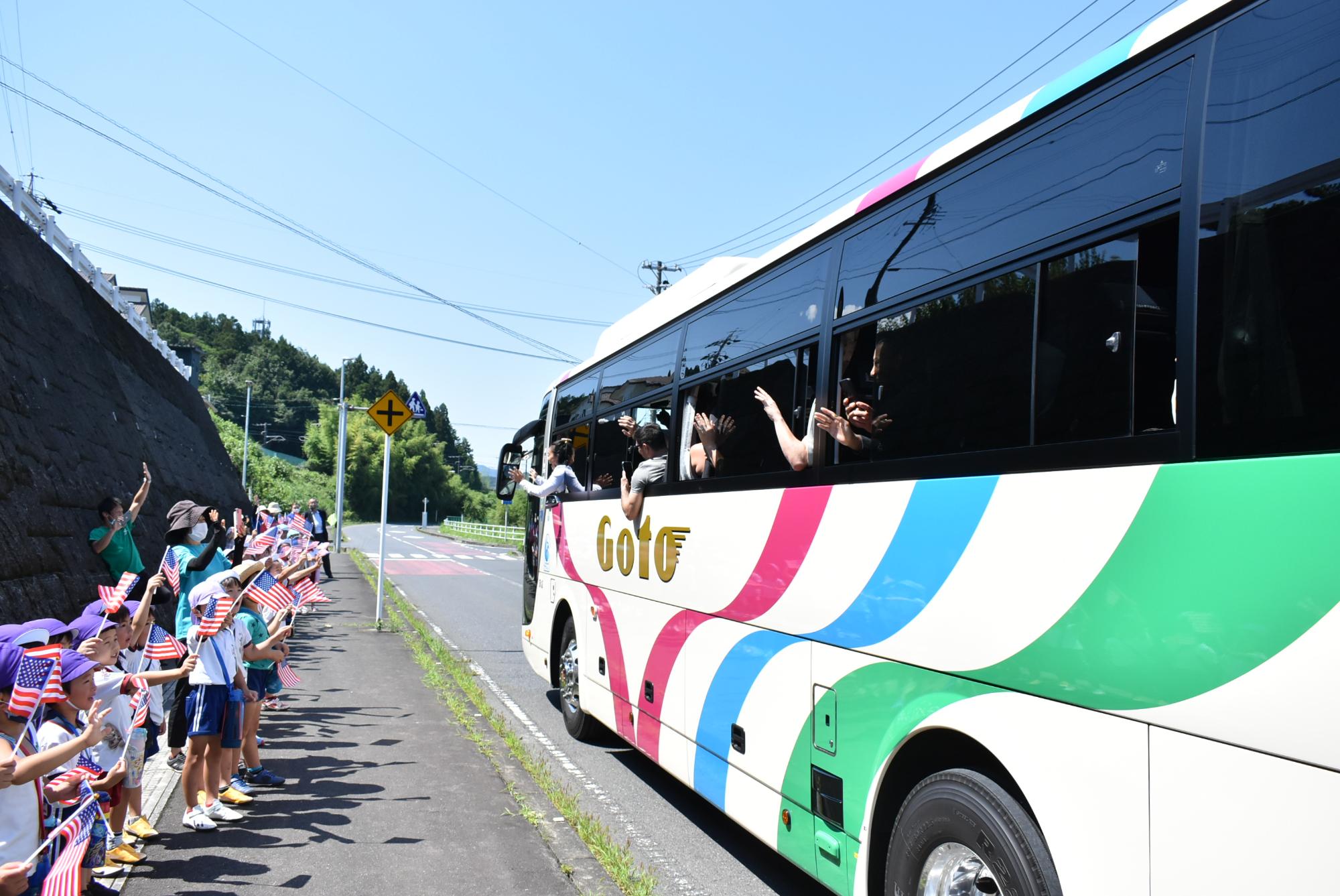 沿道で杉の子幼稚園児が並んで旗を振っています。バスが通り過ぎていきます。