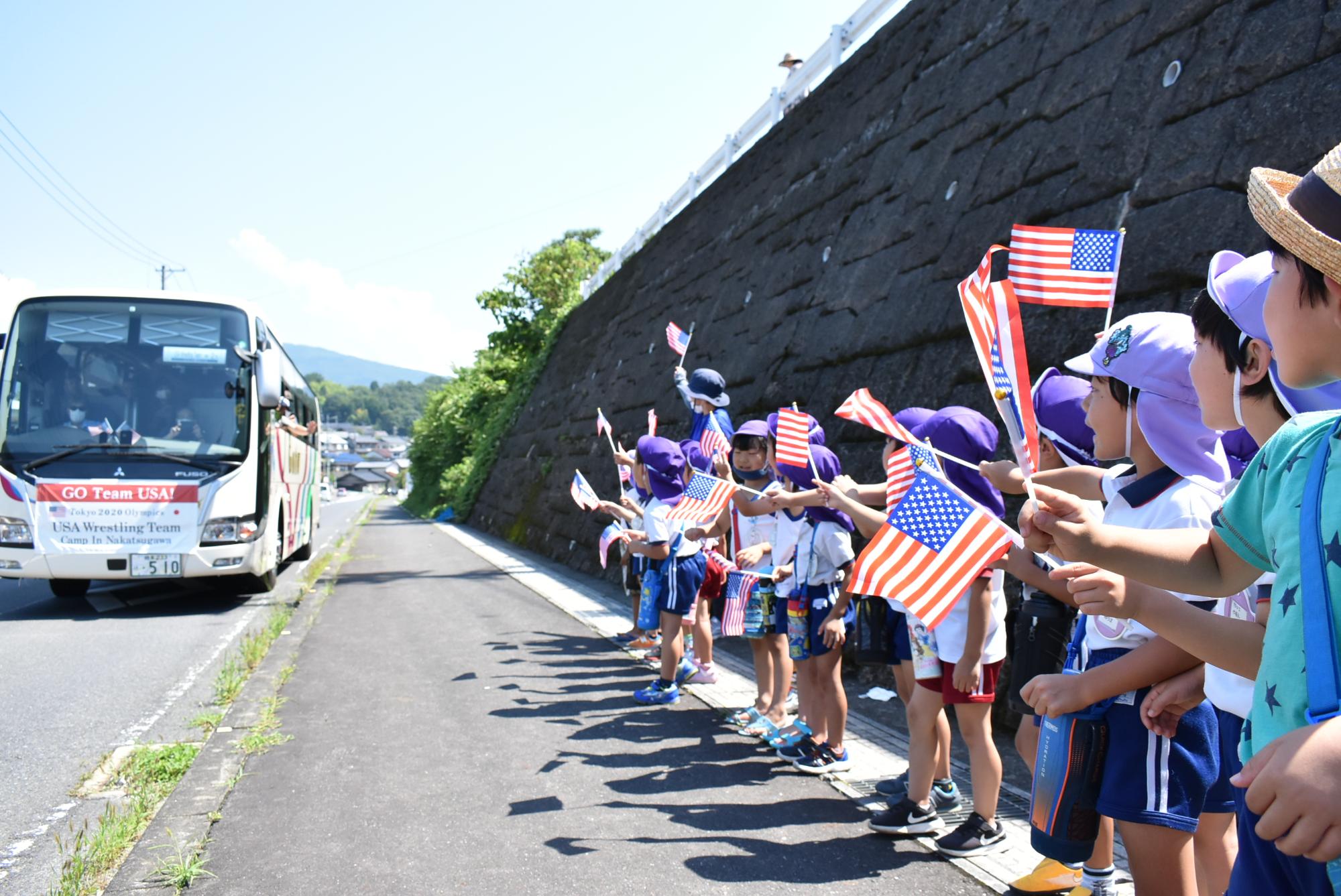 沿道で杉の子幼稚園児が並んで旗を振っています。バスが向かっています。
