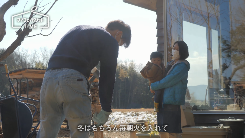 お父さんが薪の準備しているのを抱っこされた子どもとお母さんが見ています