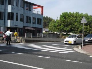 交通安全巡視活動のようす。早朝に健康福祉会館の近くの横断歩道で、センターの男性職員が交通安全の旗を使って対向車を止めて、通学中のこどもが安全に渡れるように誘導している。