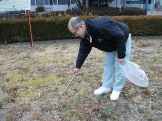 清掃活動のようす。健康福祉会館周辺の空き地で、火ばさみとごみ袋を手に持ったセンターの男性職員が、火ばさみでごみを拾い上げるところ。