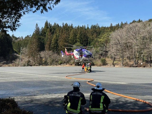 防災航空隊のヘリがバケットをつけ地面スレスレでホバリングしていることろに、地上の消防隊が放水によりバケットに補水している様子